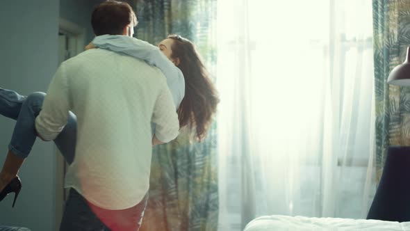 Young Couple Hugging in Hotel Room. Man Carrying Woman in Hands Indoors