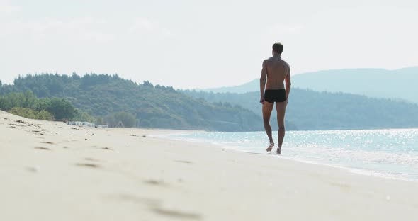 The Handsome Man with a Perfect Athletic Body in Swimming Trunks Having Fun on a Deserted Beach in