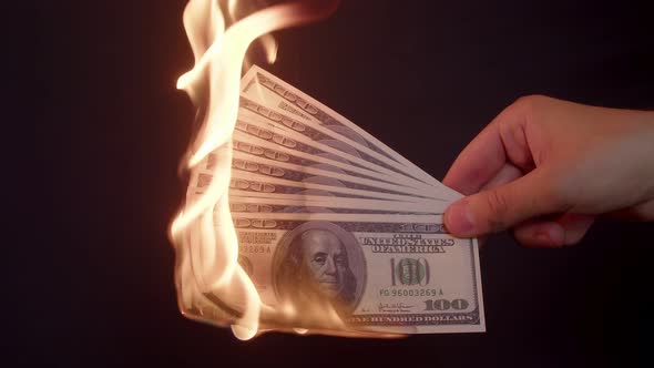 Several hundred-dollar bills in hand, which are slowly burning on a black background in close-up.