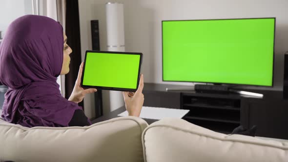 A Muslim Woman Looks at a Tablet (Horizontal) and TV (Both Green Screen) As She Sits on a Sofa