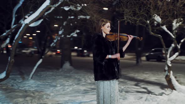 A gorgeous girl in a white dress with sparkles and in a fur coat is standing in the park