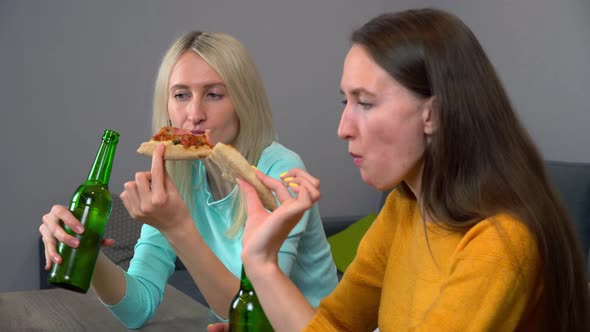 Two best happy girlfriends at home in kitchen eating pizza and drinking beer.