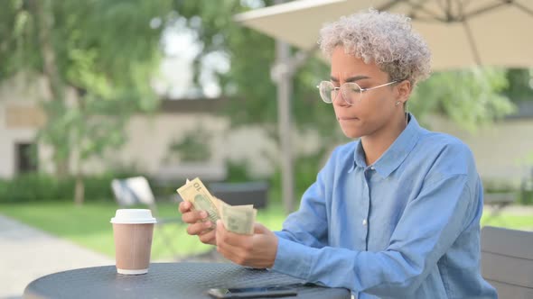 Young African Woman with Coffee Counting Dollars in Outdoor Cafe