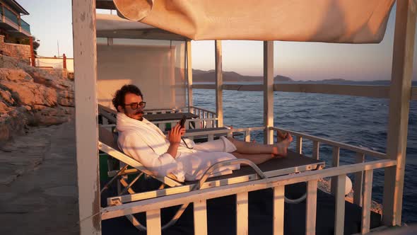 Man is Lying on Sunbed Under Tent on Coastline with Smartphone in Hands and Watching Sunset