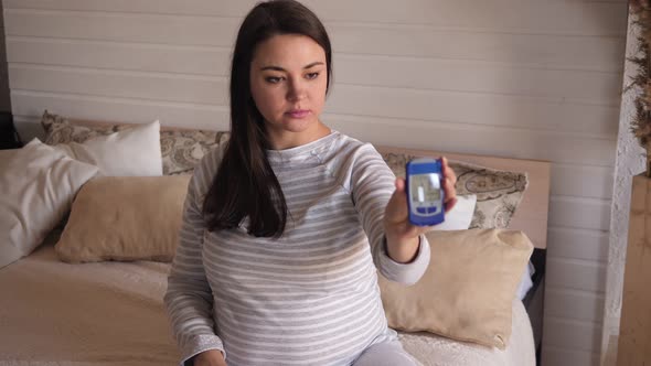 Portrait of a Pregnant Woman with a Blood Glucose Meter in Her Hands on the Bed.