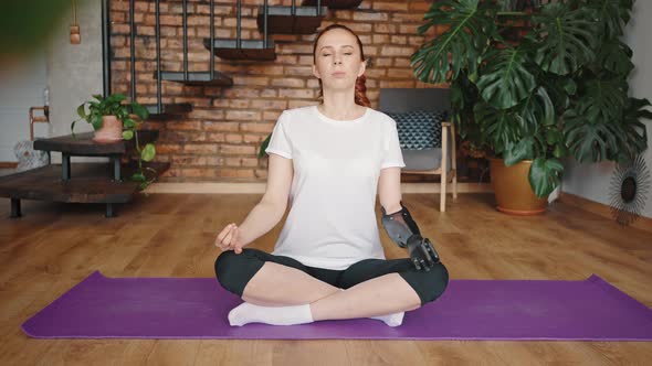 Young Adult Woman with a Robotic Hand Sits in a Lotus Position on a Yoga Mat