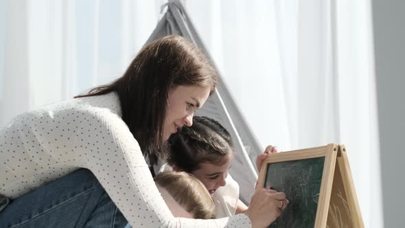 A Mother Draws with Chalk on an Easel Together with Her Little Son and Daughter