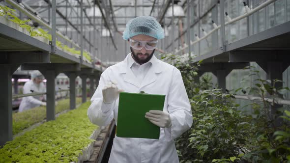 Young Confident Caucasian Man in Protective Workwear Writing Down, Looking at Camera and Smiling