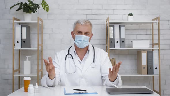 Doctor Wearing Face Mask Talking To Camera Sitting In Clinic