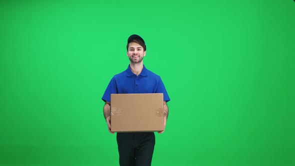 Cheerful Man Courier in Uniform Walks with a Cardboard Box in His Hands on a Green Background