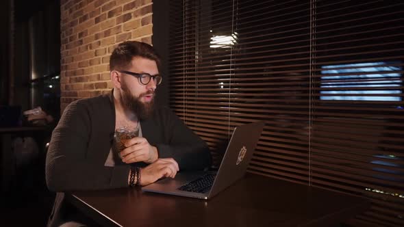 Alone Trendy Young Man Is Sitting in Cafe with Notebook and Drinking Whiskey