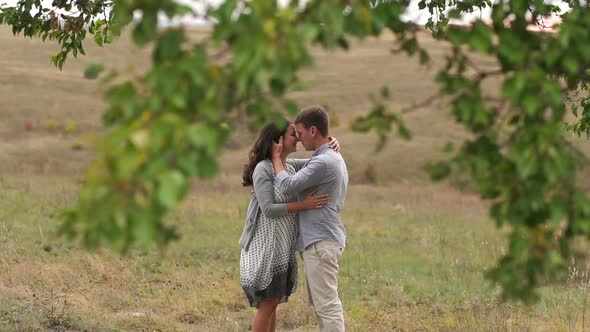 Happy Pregnant Woman with Her Husband in a Field Near a Big Tree at Sunset