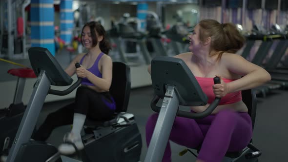 Happy Plussize Caucasian Woman Cycling Exercise Bike with Slim Young Lady Working Out at Background