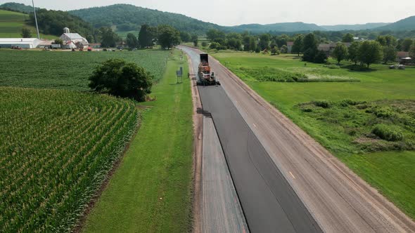 Drone view of rural highway with asphalt paver working on roadway. Green farm fields and mountains.