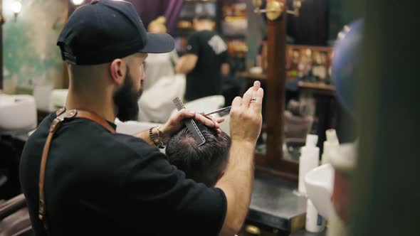 Back View of the Stylish Bearded Barber in a Black Cap Wearing Leather Apron Performing a Haircut