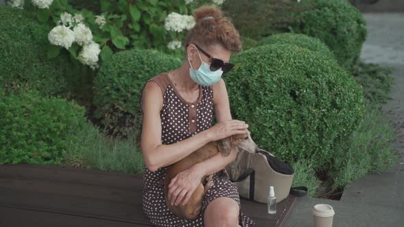 A Lone Senior Woman in Mask Spends Time with Companion Dog Sitting on Park Bench Near Home Use