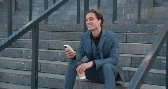 Smiling Handsome Businessman with Coffee in His Hands Sits on the Steps Near the Office Building and