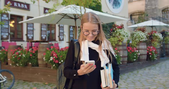 Young Woman in Glasses which Using Her Phone while Standing Near Summer Outdoors Cafe