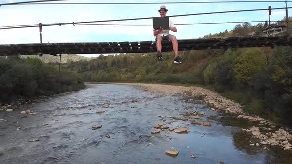 Aerial Drone View of Young Man in White Shirt Sitting on Wooden Bridge Over Mountains River and