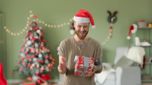 Young Handsome Bearded Tattooed Guy in Green T-shirt with Red Christmas Hat Is Taking a Gift at Home
