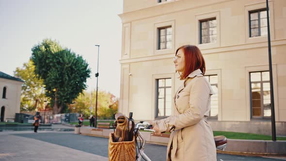 Young Hipster Redhaired Woman is Walking By City Boulevard with Her Cruiser Bike and Backpack in