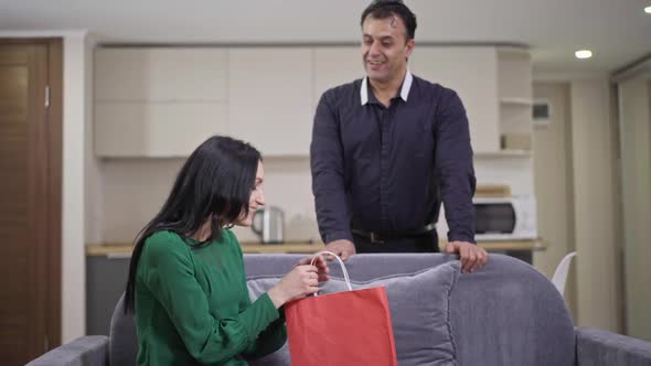 Smiling Wealthy Middle Eastern Man Giving Gift to Surprised Excited Caucasian Woman Sitting on Couch