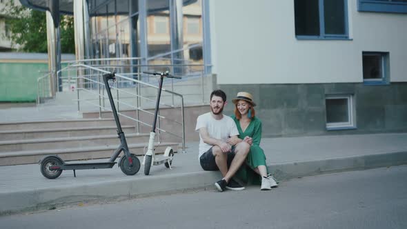 Cheerful Couple in Love Sitting on Pavement at Electric Scooters and Having Fun