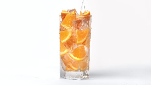 Pouring Sweet Carbonated Water Into Transparent Glass with Orange Slices and Ice Cubes Slow Motion