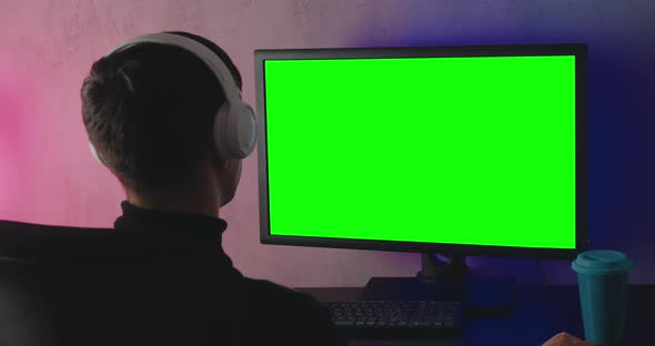 Rear View of a Young Man with Headphones Watching at Monitor with Green Screen Chroma Key Indoors