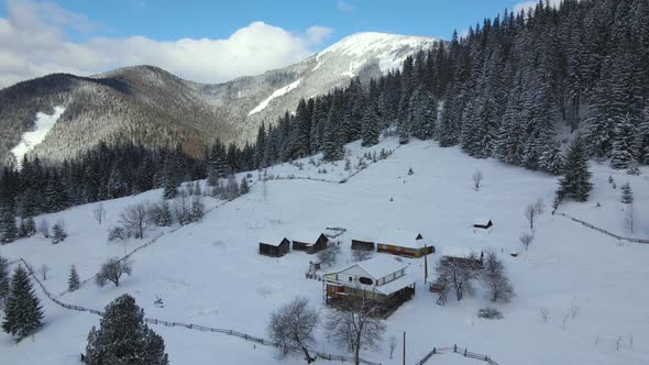 Aerial winter landscape with small rural houses between snow covered forest in cold mountains.