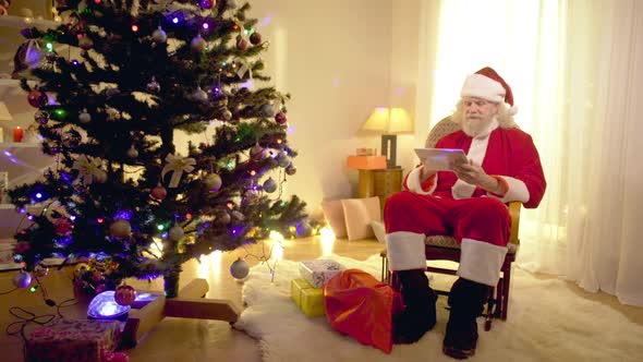 Happy Smiling Santa Clause Surfing Internet on Tablet Sitting in Comfortable Rocking Chair in Living