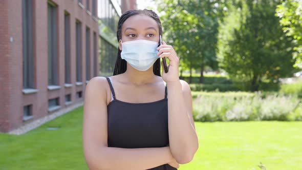 A Young Black Woman in a Face Mask Talks on a Smartphone - an Office Building and Green Park