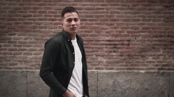 Hispanic Young Man Looking at the Camera and Posing with Hands in Pockets in Front of a Brick Wall