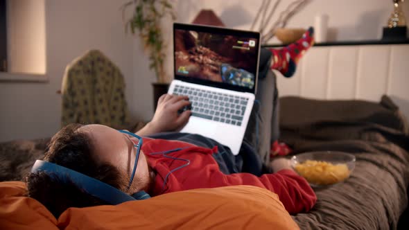 Young Man Lying on the Bed Playing Shooter Game on the Laptop