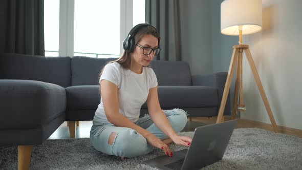 Casually Dressed Woman with Headphones is Sitting on Carpet with Laptop and Working in Cozy Room