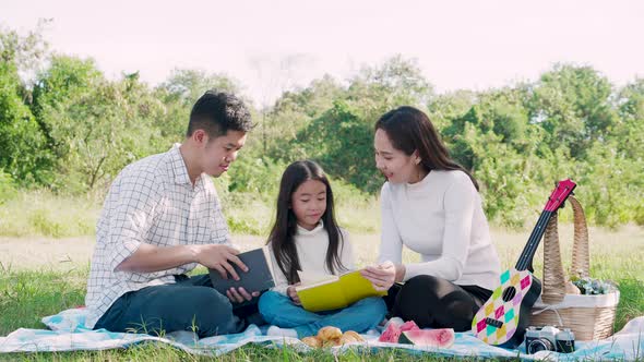 Happy family picnic. Asian parents (Father, Mother) and little girl reading a book and have fun