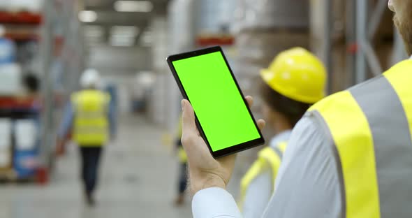 Back View of Male Worker in Warehouse Wearing Safety Vest Standing Near Shelf Using Cellphone