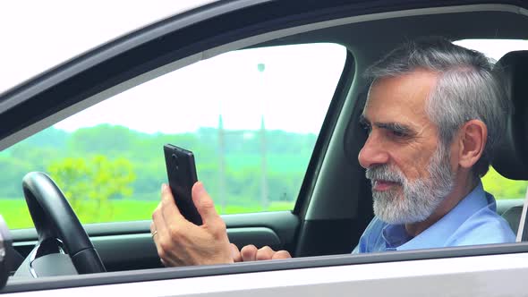 Senior Man Sits in the Car and Works on the Smartphone - Closeup Shot From Side - Countryside