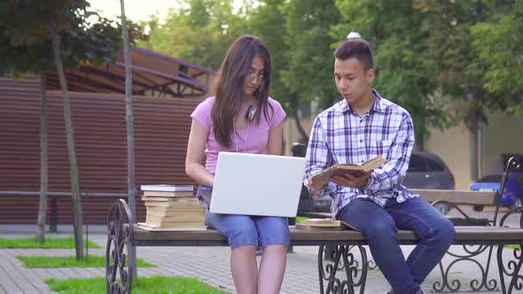 Young Asian Students Man with a Book and a Woman with a Laptop Sitting on a Bench Slow Mo