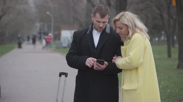 Adult Caucasian Couple Standing in Park with Suitcase and Using Smartphone. Male and Female Tourists