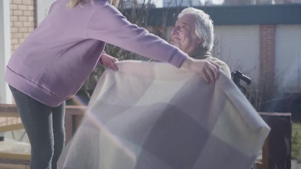 Young Unrecognizable Female Invalid Tender Covering Paralyzed Legs of Old Caucasian Man with Blanket