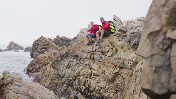 Senior hiker couple with backpacks sitting and talking on the rocks while hiking near sea shore.