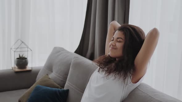 Happy African American Millennial Woman Sit and Rest on Sofa Hold Hands Behind Head