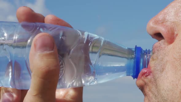 A Man Drinks Water From a Transparent Plastic Bottle Against a Blue Sky