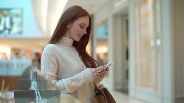 Tracking Shot of Smiling Redhead Female Using Smartphone Standing in Hall of Shopping Mall Holding