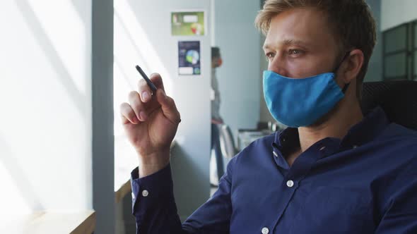 Thoughtful caucasian man wearing face mask holding pen sitting on his desk at modern office