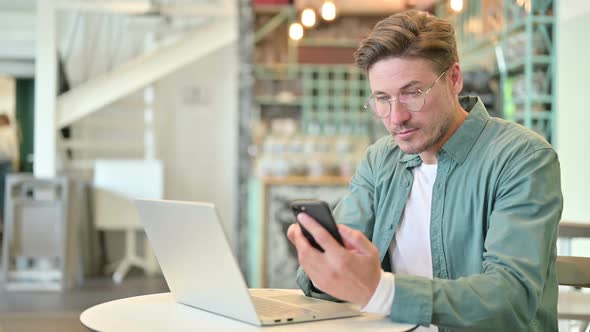 Smartphone Use By Middle Aged Man with Laptop in Cafe