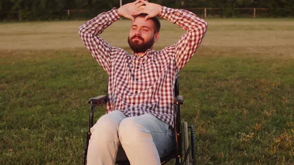 Disabled Person Man in Wheelchair on the Lawn in Rays of Setting Sun Holds His Hands Above His Head