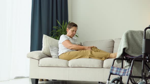 Senior Woman Massaging Her Legs While Sitting on the Couch Near the Wheelchair