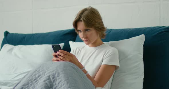 Young Woman Lying in Bed, Texting in Her Smartphone